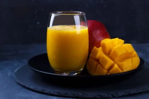 Frischer Mangosaft in einem Glas, daneben frische aufgeschnittene Mango auf einem schwarzen Teller serviert mit dunklem Hintergrund