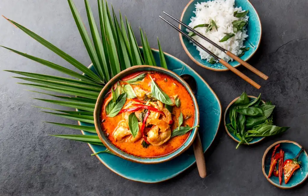thailändisches Gericht Rotes Curry mit Shrimps in kleiner Schüssel mit Palmenblatt unterlegt sowie kleiner Reisschüssel mit weißem Jasmin-Reis daneben stehend