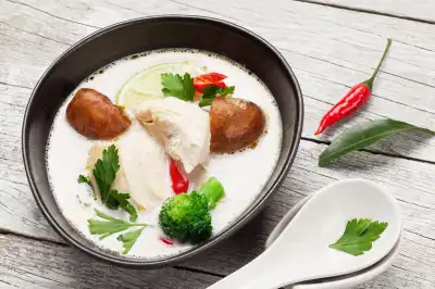 Thailändische Kokus-Suppe Tom-Kha in schwarzer Schüssel und weißem Porzellanlöffel und einer Chili auf hellem Holztisch